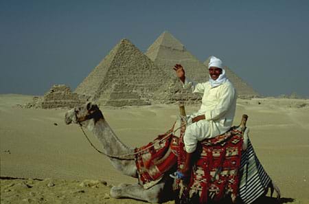 Photograph of a man on a camel in front of five pyramids in a desert.