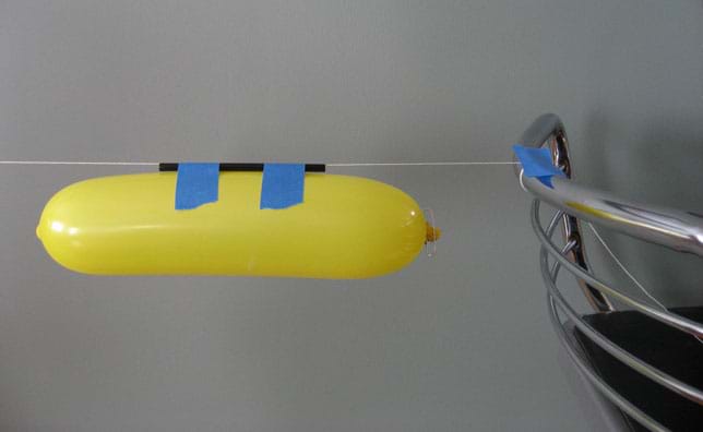 Photo shows an inflated long balloon taped to a drinking straw that hangs from a taut string attached to the back of a chair.