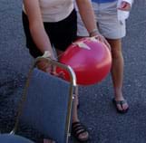 Photo shows hands holding an inflated balloon taped to a drinking straw that hangs from a taut string attached to the back of a chair.
