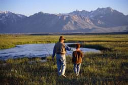 Photo shows a man and boy fishing in a field by a pond with mountain and sky in the background.