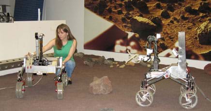 A woman stoops to make an adjustment on one of a pair of four-wheeled metal robots, about the size of large tricycles, in a large indoor sandbox.