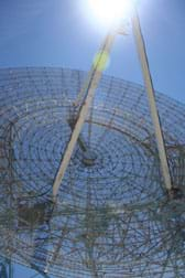 Sun shines on a huge fan-shaped apparatus made of intricate wiring, with a tripod of three metal beams at its center, pointed towards the sky.