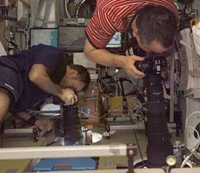 Photo shows two men pointing long-lens cameras different directions out two windows in a crowded space cabin.