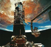 Photo shows two people in spacesuits near a tower structure in space, with the Earth overhead in the background.