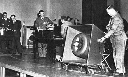 Photo shows six men in suits on a raised stage with a table of equipment.