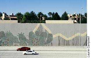 Photo shows a tall concrete wall blocking houses from cars on a multi-lane road.