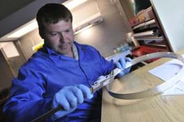 A man in a clean suit and gloves uses calipers to measure a curved piece of metal.