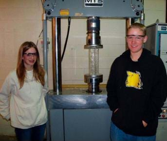 A photograph shows two students standing in front of a compression testing device with an acrylic plastic tower ready to be compressed.