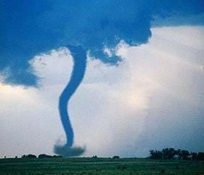 A landscape photo shows a tall and dark snaky-shaped column rising from swirling dust at its base on the ground of a flat field to masses of dark clouds high in the sky at its top.