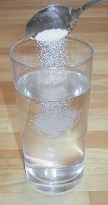 A photograph shows a spoon dropping table salt (NaCL) into a tall glass of tap water to make a saline water solution.
