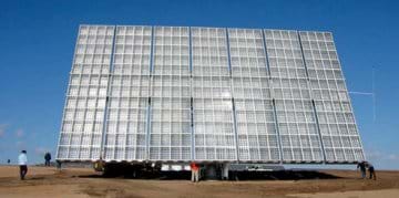 Photo shows a huge panel propped up and facing the sun in an open field. Panel is made of white and clear plastic in sliver metal grids. Panel looks very large next to the men standing near it—maybe 15 meters wide x 12 meters tall.