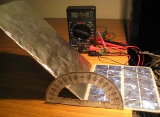 Photo shows a small photovoltaic panel with reflector panel made of foil-wrapped cardboard, hooked with wires to a multimeter.