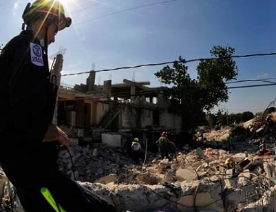 A man in a safety helmet looks into a pit and field of crumbled rubble from destroyed buildings.