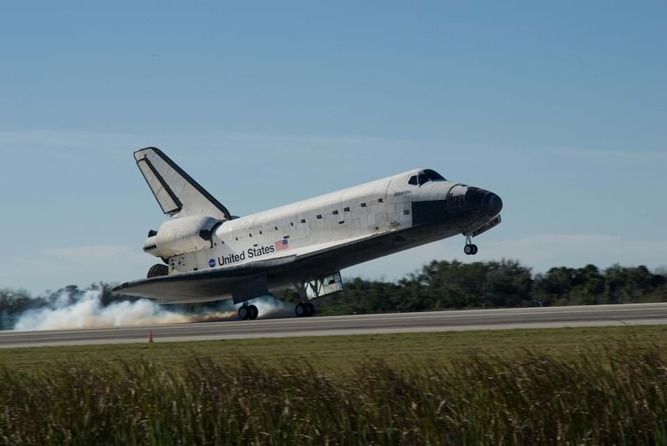 Photo shows a US space shuttle landing, with its rear wheels about to touch the runway and its nose and front wheels still in the air.