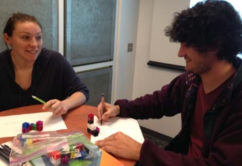 A photograph shows two teens at a table drawing two-dimensional illustrations on paper of the three-dimensional cubes on the table in front of them. 