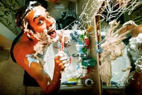 Photo shows a boy at a bathroom sink in front of a mirror. He, the sink and the mirror are covered with toothpaste, shaving cream, soap suds and silly string.