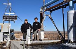A photograph shows two men standing near a water reservoir surrounded by equipment used to send water and air quality data to a remote monitoring station.