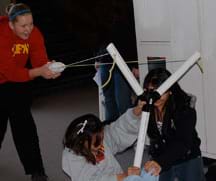 A photograph shows three students launching a folded-up t-shirt from a big y-shaped device made of PVC piping that looks like a big slingshot.