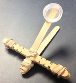 La fotografía muestra una pequeña catapulta hecha de palos de paleta, gomas elásticas y un tapòn de botella de plástico.