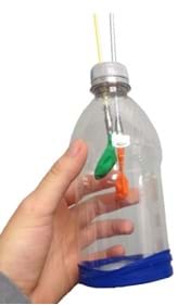 Fotografía de un modelo de pulmones que consiste de una botella de plástico y dos pajas con globos pequeños insertados en sus extremos inferiores, introducidos dentro de la botella a través del tapón.