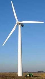 A photograph shows a white three-blade wind turbine in an open field with a tractor at its base, showing the tremendous size and height of the turbine.