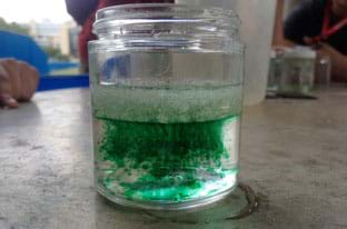 A photograph shows a transparent glass jar about halfway full of clear liquids and solids with some suspended green dye. On top of the water is a layer of mixed vegetable oil and aerogels, with the green food coloring moving downwards, away from these materials, into the water in the lower portion of the jar. 