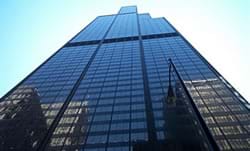 Photograph shows street view looking up at very tall glass-covered skyscraper that reflects the blue sky and nearby tall buildings.