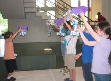 A photograph shows students standing in a circle preparing to test their disaster relief packages from the initial height, above their heads.  