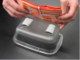 A photo shows two hands placing a fabric piece on to two rolls of duct tape placed o nthe outside bottom of a small rectangular plastic container.