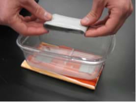 Photo shows two hands placing a long loop of duct tape into the bottom of a rectangular plastic container.