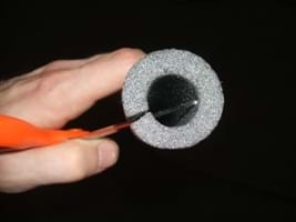 Photo shows a hand and scissors beginning to cut a tube of insulation foam.