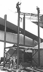 A black and white photo shows a tall I-beam structure with two men up high, working on the beams.