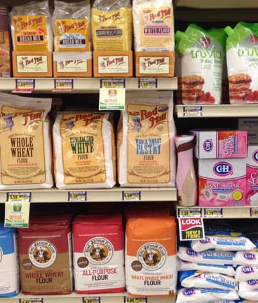 Photograph of three grocery store shelves with baking items in various containers, such as flour and grains in paper sacks, sugar in plastic bags, plastic canisters and cardboard boxes.