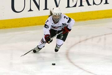 A female hockey player on ice about to hit the puck playing in the Olympics.