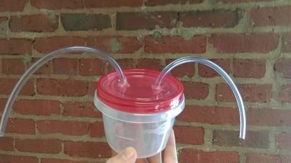 A photograph shows a hand holding a round clear plastic container with a lid. Emerging from two holes in the lid are short lengths of quarter-inch inner diameter clear plastic tubing.