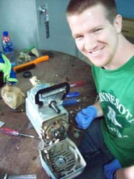 A photograph shows a smiling young man with gloves on his hands near a disassembled machine on a tabletop with parts and tools scattered nearby.