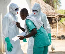 A photograph shows two healthcare workers fully gowned in white biohazard suits and blue gloves standing near a thatched-roof home as a third person writes one worker's name and time on the outside of his/her suit.