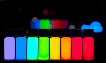 A photograph in a dark room shows a lineup of eight vivid and glowing bottles of quantum dots composing the ROYGBIV color spectrum from violet to deep red.