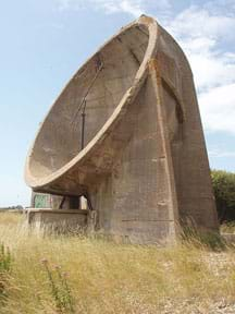 A photograph shows a sound mirror in a grassy field in England. It’s a huge concrete bowl-shaped structure pointing towards the sky.