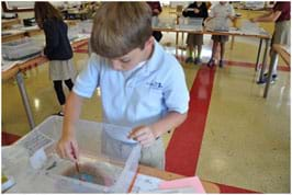 A photograph of a classroom shows a boy in the foreground using a brush to drop ink into a container of shallow water at his station, while several other students work at other workstations on tables in the background.