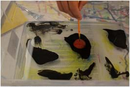 A photograph shows a paintbrush hovering over a circle of red ink enclosed by an area of black ink floating on a shallow pool of water in a clear, oblong container.