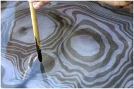 A photograph shows a bamboo paintbrush delicately poised above a shallow pool of water. On the surface of the water is a beautiful, rippling pattern of black ink concentric rings.