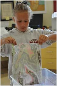 A photograph shows a girl using two hands to gently lifting a colorful piece of paper off the water surface of a rectangular tub on a table top.