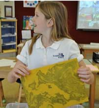 A photograph shows a girl beaming with joy as she lifts a yellow-paper print from a container of water.