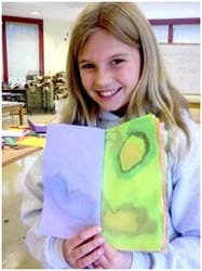 A photograph shows a girl holding a colorful packet of paper covered with water-color looking free-form ink images.