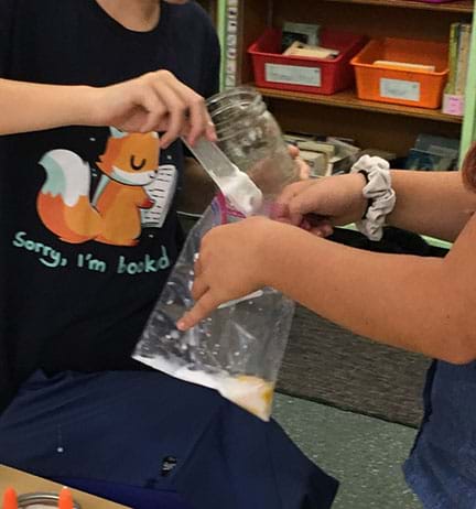 One student adds a tablespoon of borax water to a plastic bag with glue water in it being held by another student. 