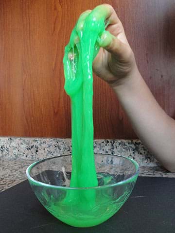 A child’s hand stretches green slime upwards from a bowl.