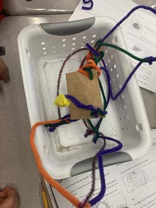 Photo shows a tangle of pipe cleaners suspending a piece of cardboard inside a white bin.