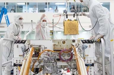 A photo showing three engineers wearing white protective cleanroom suits, gloves, and face masks working inside a laboratory to install a gold, cube-shaped instrument into the open body of a Mars rover. The rover is surrounded by exposed wiring, tubing, and mechanical components, with solar panels visible on either side. 