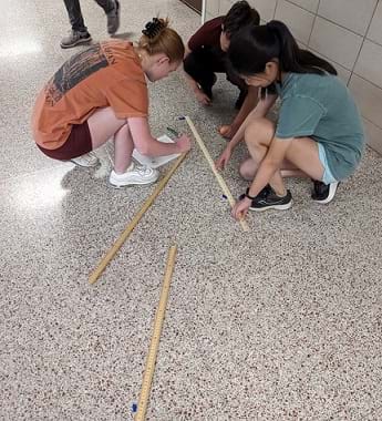 Three students crouch in a hallway, to be close to the floor. On the floor are several pieces of tape at various locations. One student holds a meter stick to measure the distance between two tape marks. A second student writes measurement values on a worksheet. A third student observes the process of the other two students. Two more meter sticks lay nearby on the floor as well.